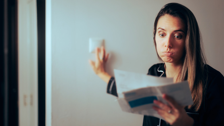 A woman looks at her utility bill while adjusting a thermostat