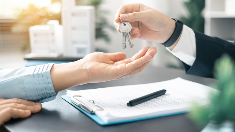 A real estate agent hands keys to a person over a desk filled with paperwork