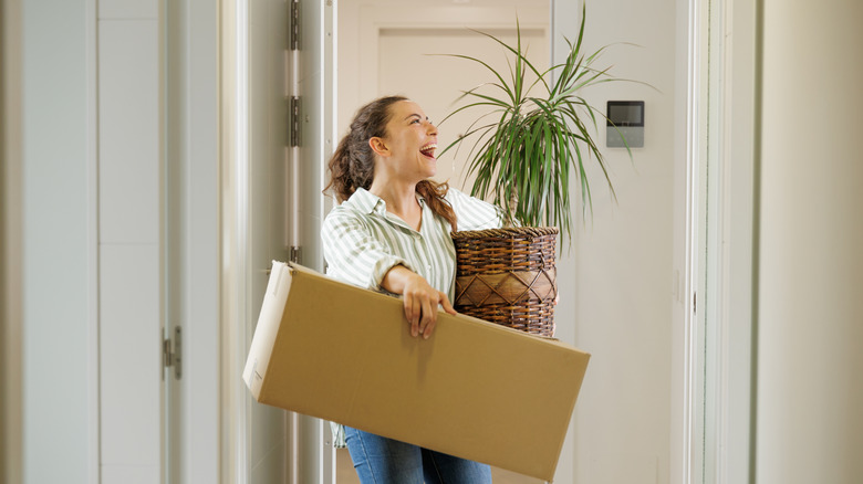 A smiling woman holds a plant and large moving box
