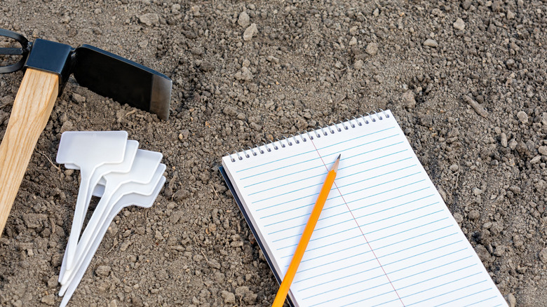 A notebook and assorted gardening tools lay on top of bare dirt.