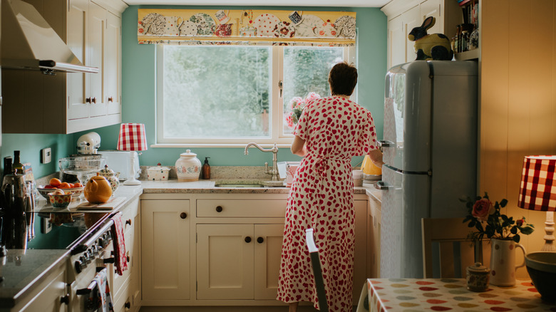 A woman washes dishes in a cramped kitchen with mismatched appliances.