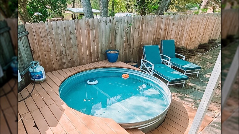 Stock tank pool in backyard with wooden deck around it and two blue lawn chairs