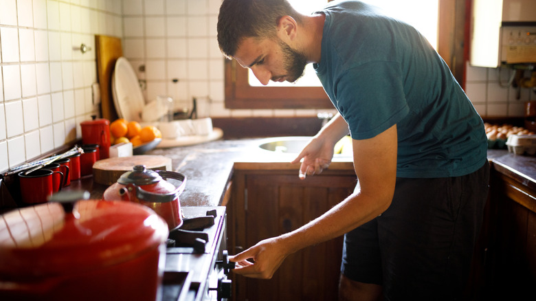 A young man turns a stove knob to turn on the flame for the kettle.