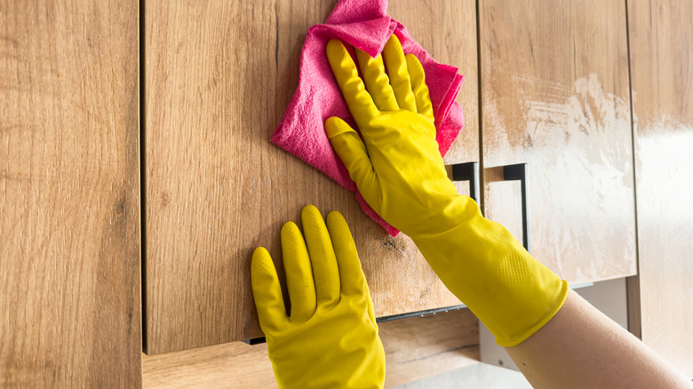 Person wears yellow rubber gloves while they apply a dish soap mix to wood cabinets with a pink towel.