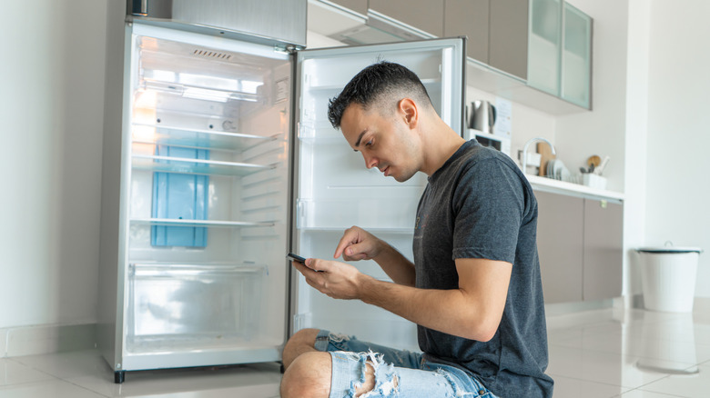 a man sitting near an open refrigerator browsing on his cell phone