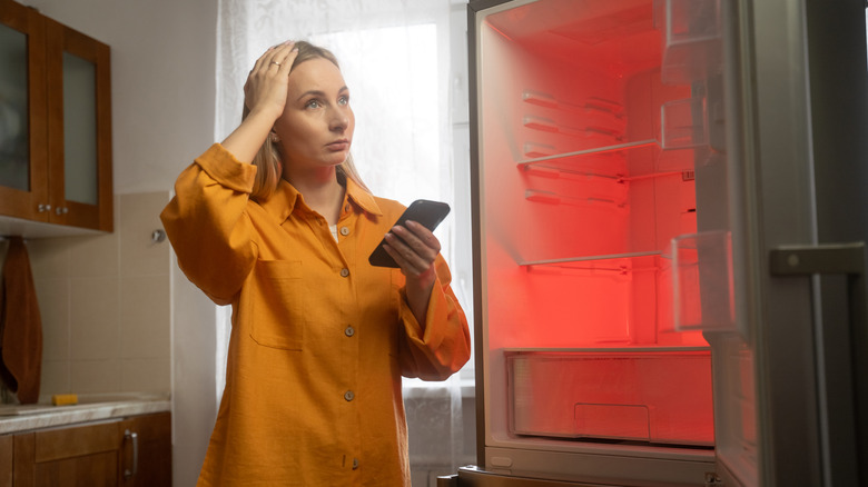 A woman trying holding her head and a phone next to an open refrigerator