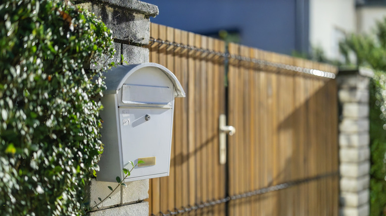A white mailbox in a brick column sandwiched between a hedge and a residential gate