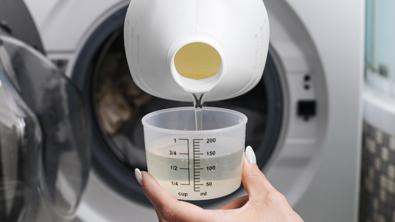 A person pouring liquid laundry detergent into a clear plastic cup in front of a washer