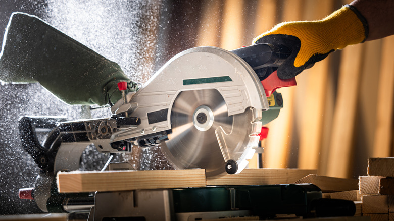 A gloved hand using a circular saw to cut a large board of wood.