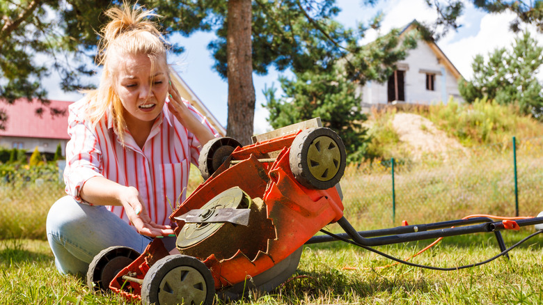 A woman sitting in the grass inspecting an overturned lawn mower, looking concerned