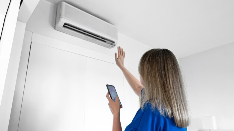 A woman uses a smart HVAC control app on their phone while feeling air movement from AC unit