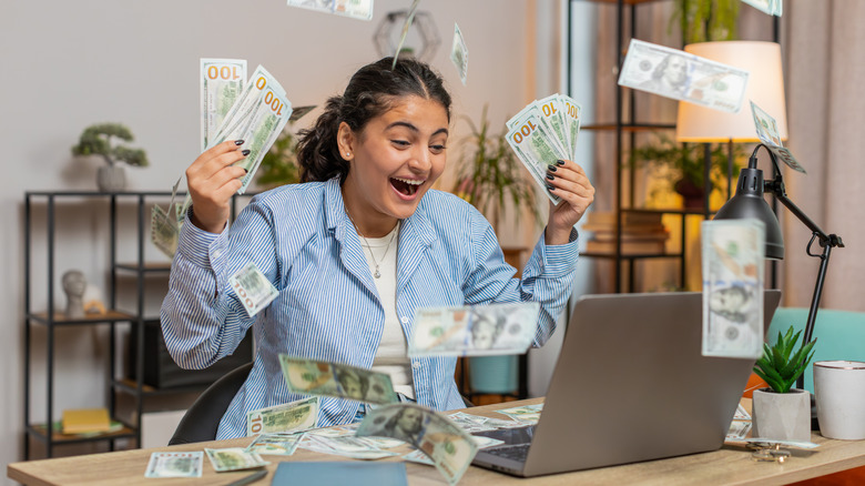 A woman throwing hundred dollars bills in the air, seated in her home office,