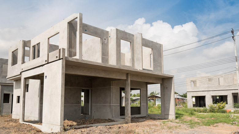 A two-story concrete house under construction, featuring an unfinished structure with open window spaces, set on a dirt lot with a clear sky and nearby building sites in the background.