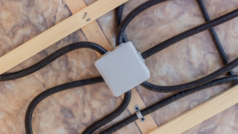 Electrical wires run through a grey junction box attached to a wooden ceiling frame stuffed with insulation