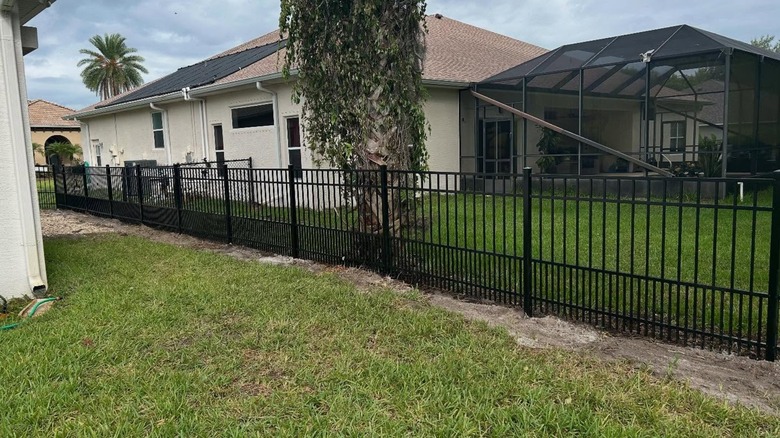 A black puppy picket fence surrounded  by green grass and palm trees with tan stucco house in the background.