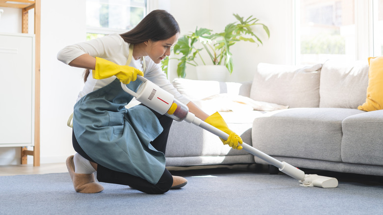 A cleaner wearing a blue apron and yellow gloves vacuuming under a couch