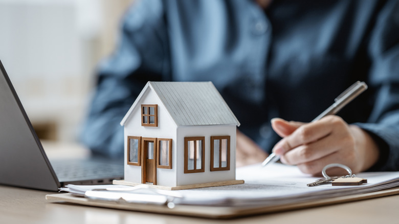 model house on a desk next to a person working on a computer and writing notes