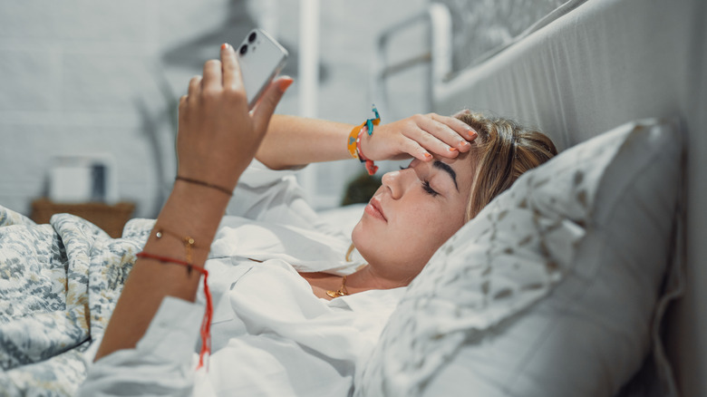 A girl holds her head in pain while using a smartphone in bed