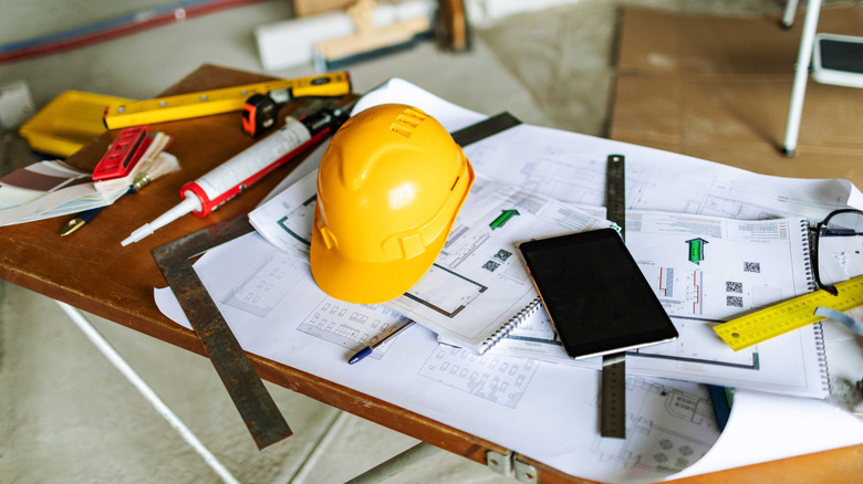 Construction tools a tablet and a hard hat sitting on a table over blueprints in an unfinished room
