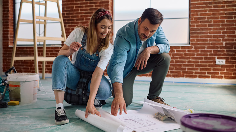 man and woman in work clothes in a brick room with a ladder and paint looking at blueprints