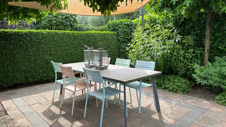 Outdoor patio with a modern table, pastel chairs, and overhead shade sail, surrounded by lush green hedges and plants.