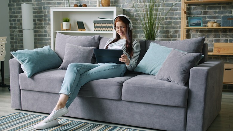 A smiling person sitting on a gray couch watching media with a tablet and headphones