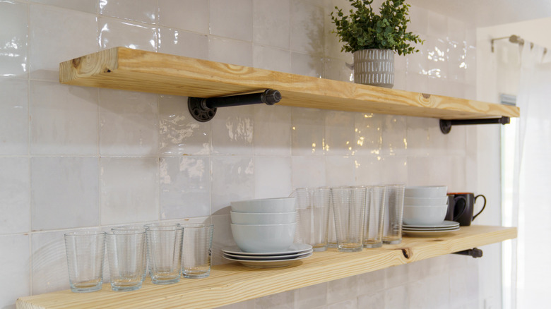 Dishes, glasses, and potted plants displayed on top of two wood shelves with industrial pipe brackets on a white tile wall.