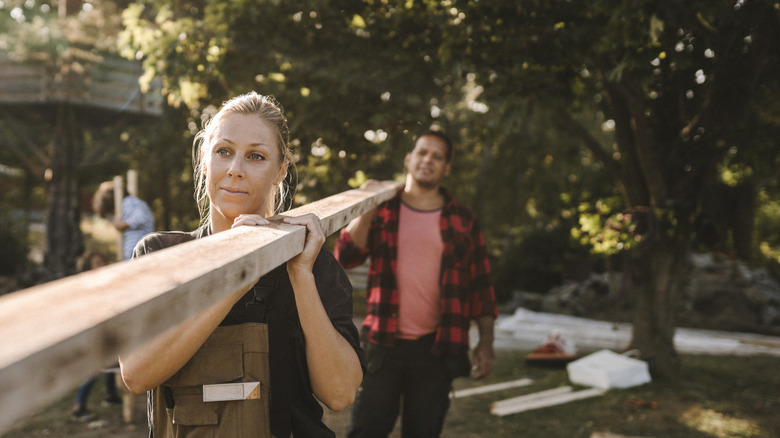 A woman carries a timber plank across a backyard undergoing renovation.