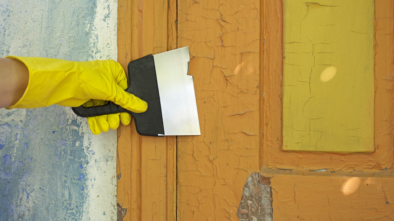 A person uses a scraper to remove alligatoring orange paint from a door