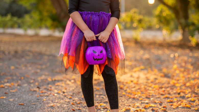 Child holding a purple Halloween bucket on a street with fallen yellow leaves