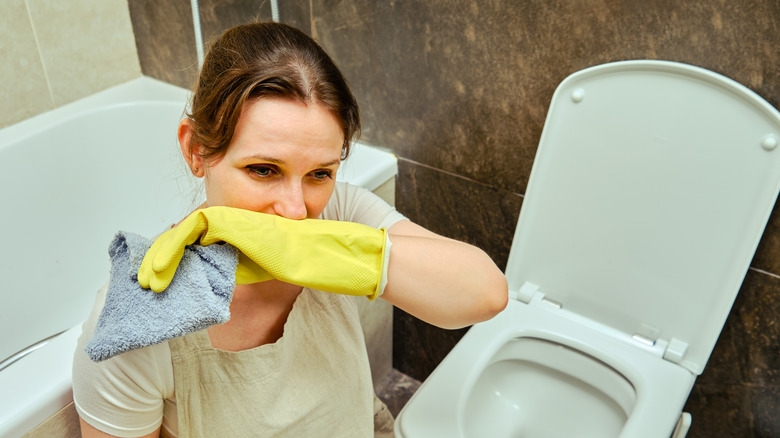 Woman crouching by toilet with cleaning gloves on, covering her nose