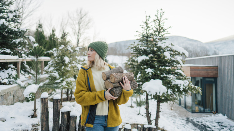 A woman in a yellow jacket and a green beanie grabbing logs from her backyard with trees while snow is on the ground and plants
