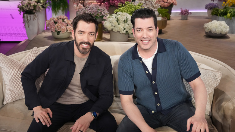 Drew and Jonathan Scott sitting on a love seat with colorful potted flowers behind them.