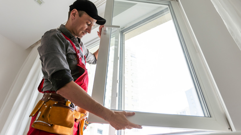 A man wearing a black baseball cap and leather toolbelt installs a casement window