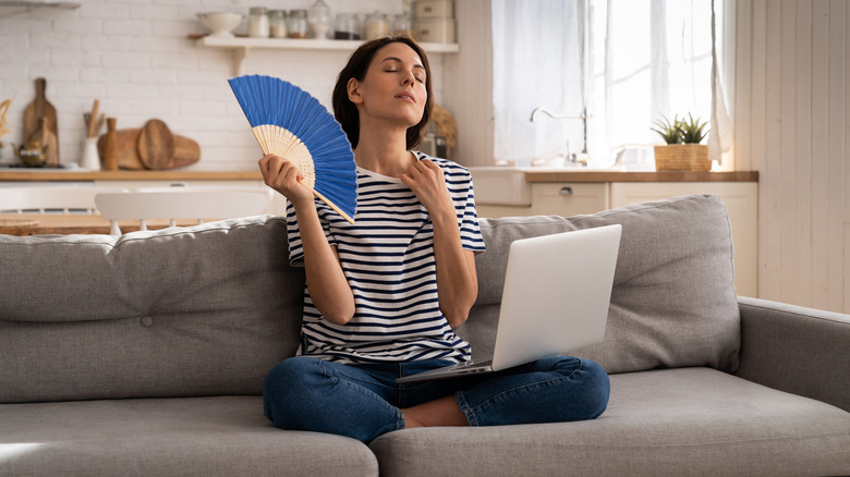 A person sits on a couch with a hand fan and laptop