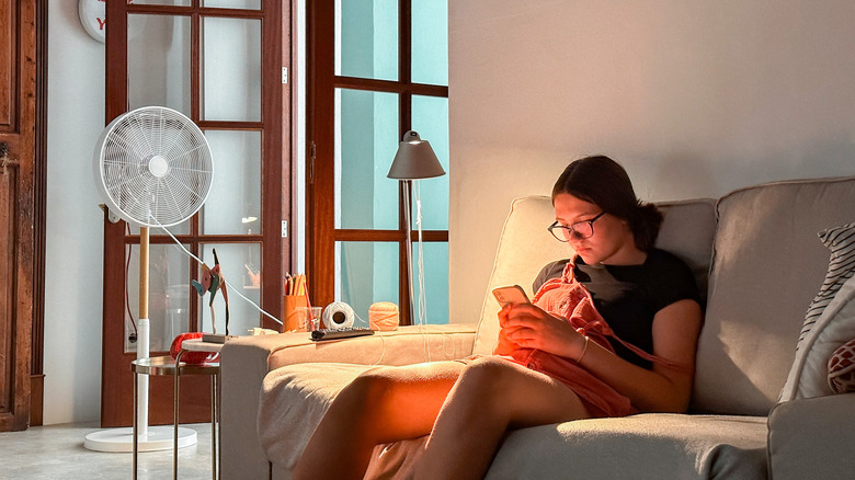 A strategically placed fan points at a person sitting on a couch in a dim room
