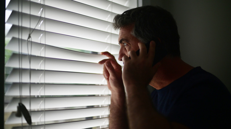 A man on the phone peeking through his blinds.
