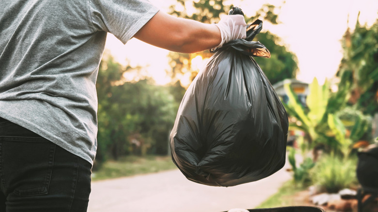 Person holding a full black trash bag to place it in the garbage bin outside