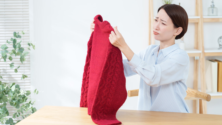 A woman is upset to find holes in her red sweater, which she is holding up