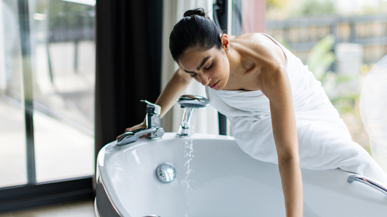Woman sitting on the edge of the tub looking in at the drain.