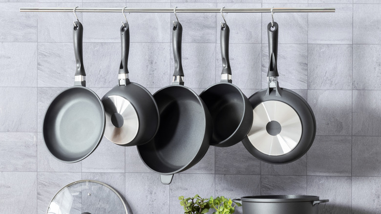 Set of cookware pans hanging via hooks from a metal rod in a modern kitchen with grey backsplash