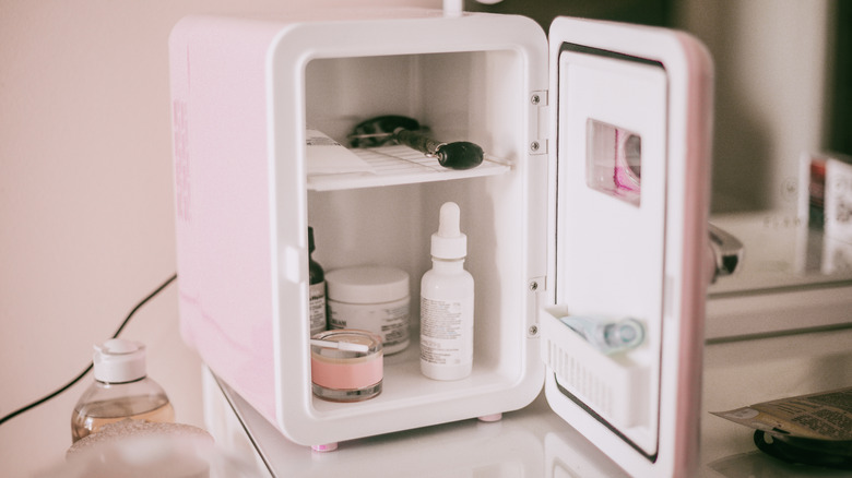 A pink mini fridge in a bathroom with its door open, showing skincare, makeup and other beauty products inside.