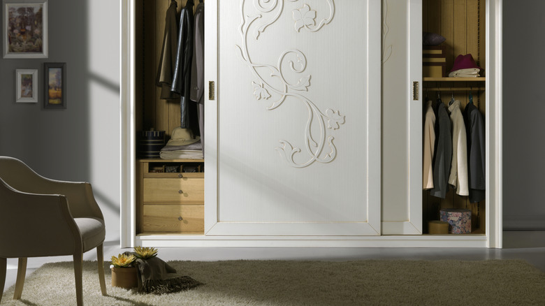 A bedroom with ornately carved white wardrobe with sliding doors and clothes hanging inside