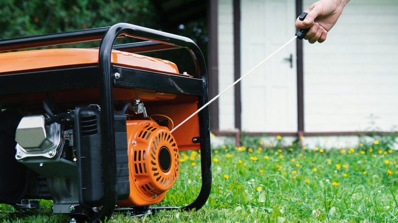 Hand starts a portable electric generator standing on the grass in front of a summer house in summer evening