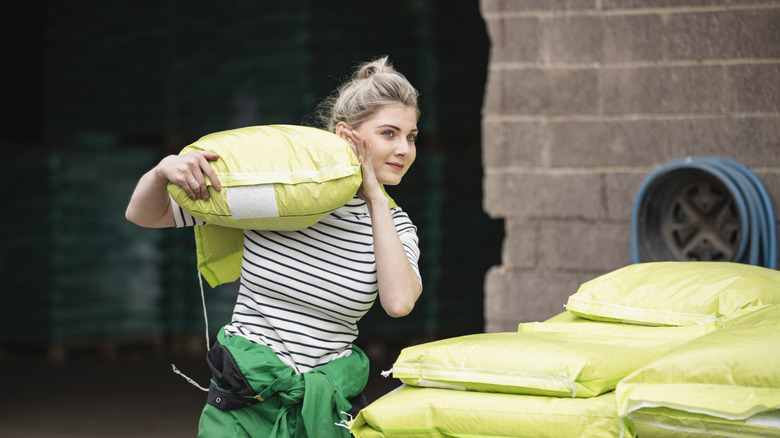 A woman carrying a yellow potting soil bag on her shoulder