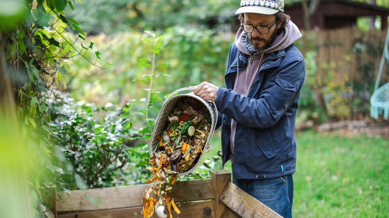 A man in a blue jacket and jeans dumps a bucket of food scraps into a large compost bin.