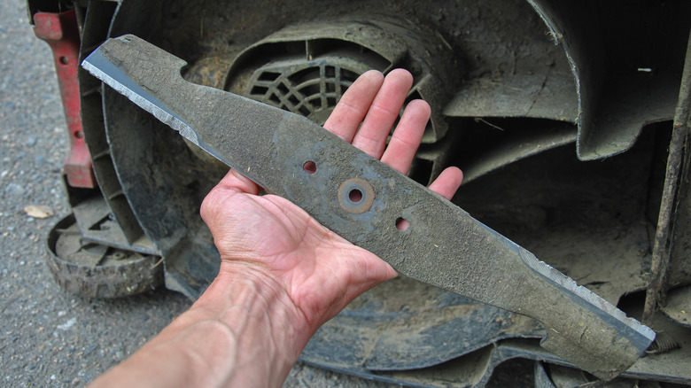 A person holding a used and dirty lawn mower blade in their hand, with the mower on its side in the background