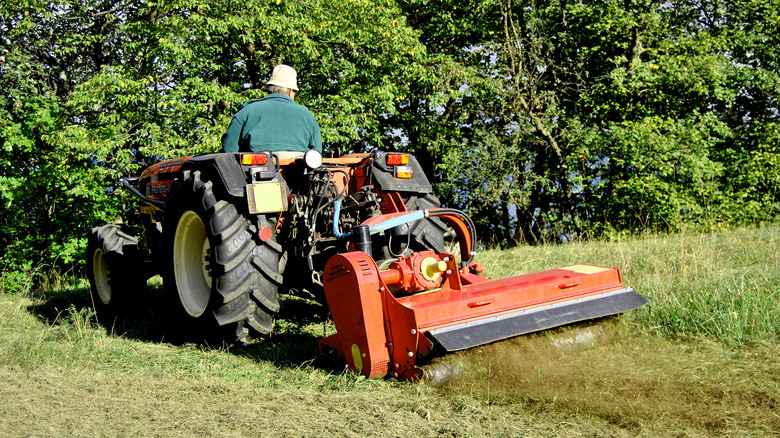 A person with a bucket hat driving a tractor and pulling a smaller bush hog mower