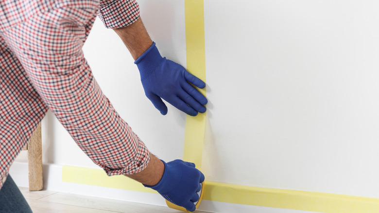 A person wearing blue rubber gloves applies yellow painter's tape to a white wall.