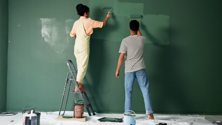 A couple paints a living room wall deep green using rollers and a step ladder.